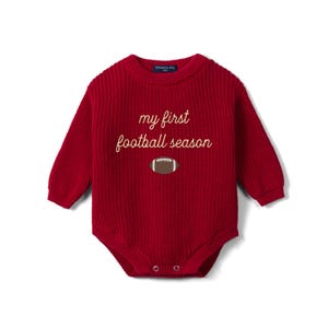 May include: A long-sleeved, deep red knitted baby bodysuit featuring a ribbed texture and a crew neckline. The chest displays the embroidered text 'my first football season' in a light cream color, positioned above a small brown football graphic.