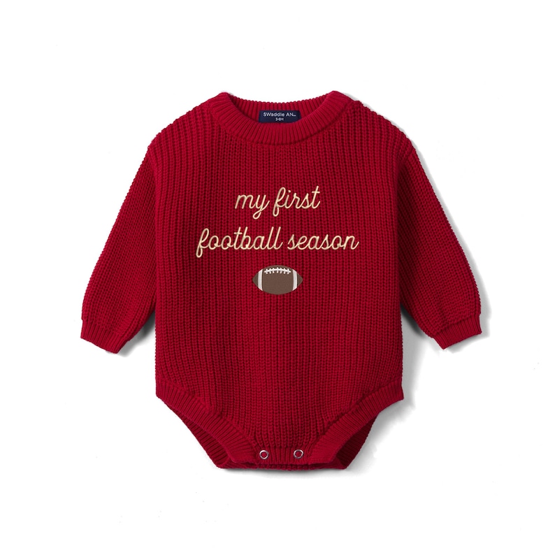 May include: A long-sleeved, deep red knitted baby bodysuit featuring a ribbed texture and a crew neckline. The chest displays the embroidered text 'my first football season' in a light cream color, positioned above a small brown football graphic.