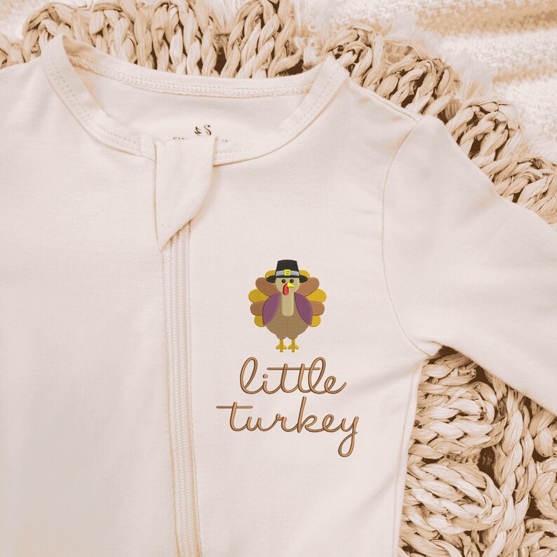 May include: Cream-colored baby onesie with a zipper closure. The onesie features an embroidered turkey wearing a pilgrim hat and the words "little turkey" in a cursive font. The background is a woven, beige surface.