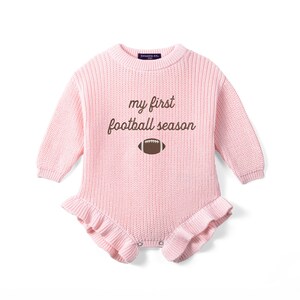 May include: A light pink long-sleeve knit romper featuring a crew neckline and ruffled leg openings. The chest displays the brown embroidered text "my first football season" above a small brown and white football graphic icon.