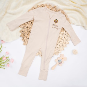 May include: A beige baby sleeper with a full-length zipper and the words "little turkey" printed on the front. The sleeper features a cartoon turkey graphic. A wooden rattle and a small ball are also visible.