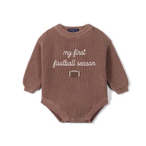 May include: A long-sleeved, brown, chunky knit baby bodysuit featuring a crew neckline and ribbed cuffs. The chest displays the white embroidered text 'my first football season' positioned above a small, brown and white football graphic.