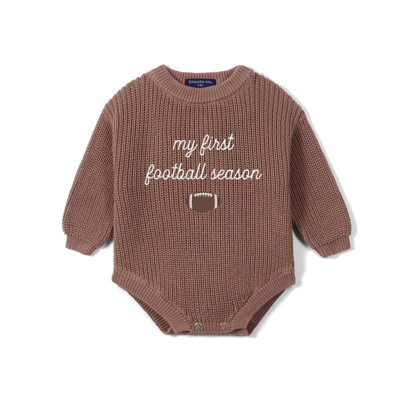 May include: A long-sleeved, brown, chunky knit baby bodysuit featuring a crew neckline and ribbed cuffs. The chest displays the white embroidered text 'my first football season' positioned above a small, brown and white football graphic.