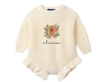 May include: A cream-colored long-sleeve knit romper featuring a gingerbread man graphic surrounded by a wreath of green leaves and red berries. The name olivia is printed in dark green script below the gingerbread man design.