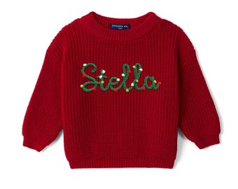 May include: A deep red long-sleeve knit sweater featuring a ribbed crew neckline and cuffs. The front displays the name Stella embroidered in green yarn, accented with small, colorful pom-poms resembling holiday lights. A tag reads Swaddle AN.