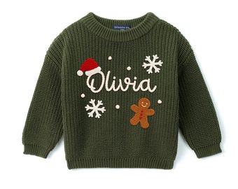 May include: An olive green knit sweater featuring the name Olivia embroidered in white script. The design includes a red and white Santa hat, two white snowflakes, and a brown gingerbread man with red buttons on the front of the garment.