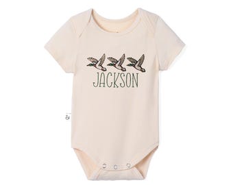 May include: A cream-colored short-sleeve bodysuit featuring a graphic of three flying mallard ducks in a row above the name JACKSON printed in dark green capital letters. The garment has a round neckline and three metal snaps at the bottom.