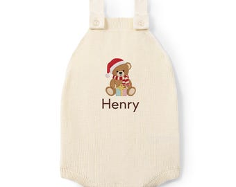 May include: An off-white knitted romper featuring two shoulder straps with buttons. The center displays an embroidered brown teddy bear wearing a red and white Santa hat and scarf, holding a gift box. The name Henry is embroidered below.