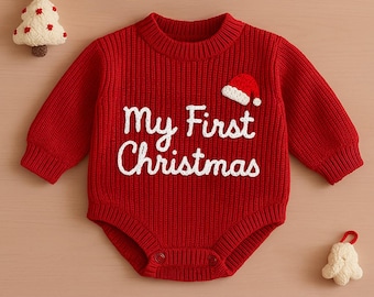 Puede incluir: Body rojo de punto para bebé con el texto "My First Christmas" en blanco. Un pequeño gorro de Papá Noel está bordado en el pecho. El body tiene mangas largas y cierres de presión. Un pequeño árbol de Navidad y una campana están en el fondo.