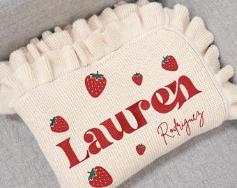 Può includere: Coperta per neonati in maglia color crema con fragole ricamate in rosso e il nome "Lauren Rodriguez". La coperta ha un bordo a balze e poggia su una superficie grigia.