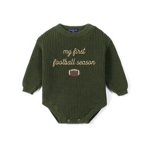 May include: An olive green long-sleeve knit romper featuring a crew neckline and ribbed texture. The chest displays the embroidered text "my first football season" in a light tan script font, positioned above a brown and white football graphic.