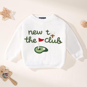 K&ouml;nnte beinhalten: Wei&szlig;er Strickpullover mit dem gr&uuml;nen Text "new to the club", mit einem Golfball, einer Fahne und Gr&uuml;n. Der Pullover hat lange &Auml;rmel und einen Rundhalsausschnitt. Ein lustiges, sportliches Design f&uuml;r Golfbegeisterte.