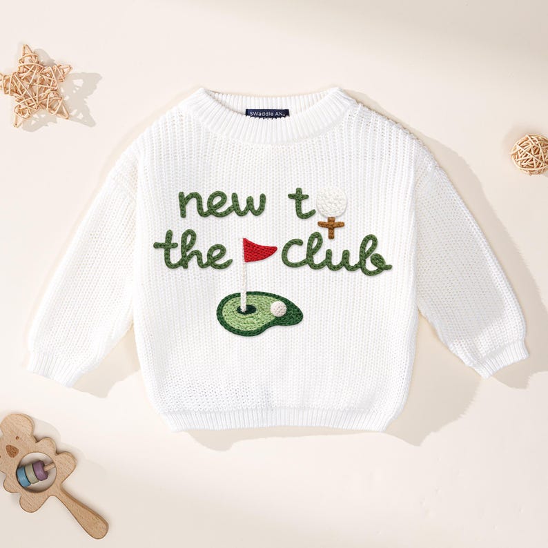 K&ouml;nnte beinhalten: Wei&szlig;er Strickpullover mit dem gr&uuml;nen Text "new to the club", mit einem Golfball, einer Fahne und Gr&uuml;n. Der Pullover hat lange &Auml;rmel und einen Rundhalsausschnitt. Ein lustiges, sportliches Design f&uuml;r Golfbegeisterte.