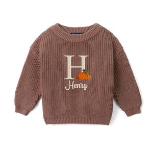 May include: A mauve long-sleeve knit sweater featuring a large cream-colored letter H embroidered on the front. Next to the letter is an embroidered orange pumpkin with green leaves and the name Henry stitched in cream cursive script below it.