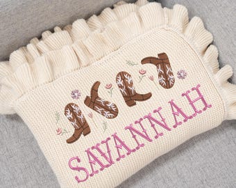 May include: Cream-colored knitted baby blanket with a ruffled edge. The blanket features embroidered brown cowboy boots, small flowers, and the name "SAVANNAH" in pink lettering. The blanket is on a gray surface.