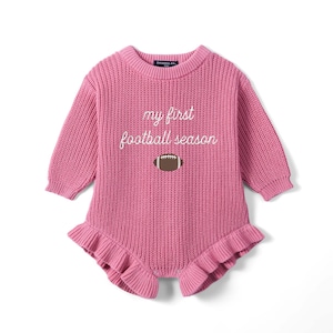 May include: A long-sleeved, pink knitted romper featuring a ribbed crew neckline and cuffs. The chest displays the white embroidered text 'my first football season' above a brown and white football graphic. The leg openings have ruffled edges.
