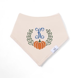May include: A cream-coloured triangular bandana bib featuring an embroidered design of a blue letter K centred above an orange pumpkin, flanked by two green leafy branches. A small white tag with a swan logo and text is attached to the side.