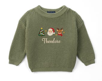 May include: An olive green chunky knit sweater featuring a crew neckline and long sleeves. The center chest displays embroidered icons of a Christmas tree, Santa Claus, and a reindeer, with the name Theodore stitched in white cursive text.