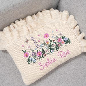 May include: Cream-colored knitted baby blanket with a floral embroidery design. The blanket features pink, purple, and blue flowers, green leaves, and a butterfly. The name "Sophia Rae" is embroidered in pink script.