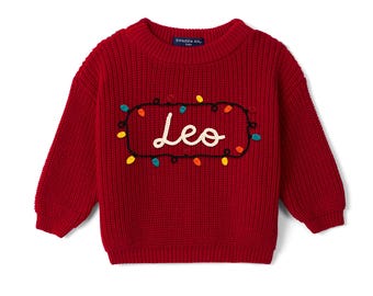 May include: A deep red long-sleeve knit sweater featuring a crew neckline and ribbed cuffs. The center displays the name Leo embroidered in white script, surrounded by a black embroidered string of colorful Christmas light bulbs.