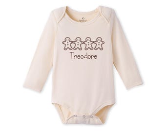 May include: A long-sleeve cream-colored baby bodysuit featuring a brown graphic of four gingerbread people holding hands. Below the gingerbread figures, the name Theodore is printed in a brown, rounded, handwritten-style font.