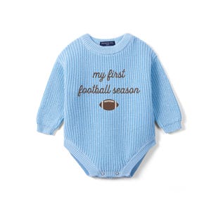 May include: A light blue long-sleeve knit bodysuit featuring a crew neckline and ribbed cuffs. The chest displays the brown embroidered text "my first football season" above a brown and white football graphic. The garment has a snap closure at the base.