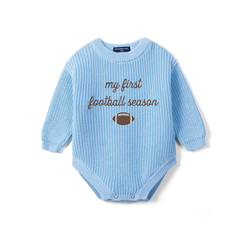 May include: A light blue long-sleeve knit bodysuit featuring a crew neckline and ribbed cuffs. The chest displays the brown embroidered text "my first football season" above a brown and white football graphic. The garment has a snap closure at the base.