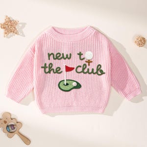 K&ouml;nnte beinhalten: Ein hellrosa Strickpullover mit dem Schriftzug "new to the club" in gr&uuml;ner Schrift. Der Pullover hat ein Golf-Motiv mit Golfball, Fahne und Gr&uuml;n. Der Pullover hat einen Rundhalsausschnitt.