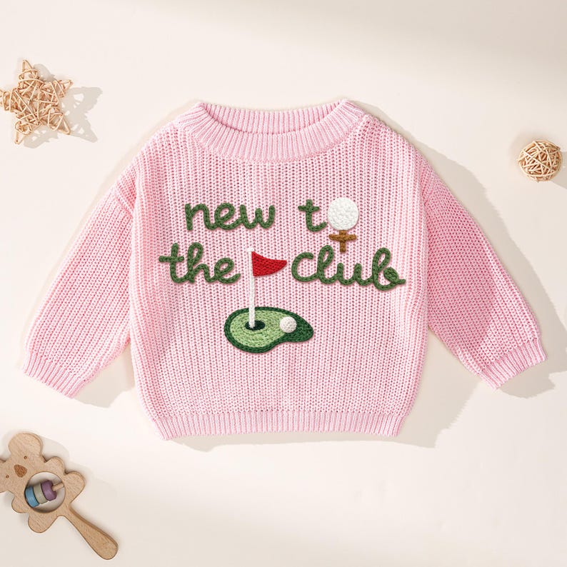 K&ouml;nnte beinhalten: Ein hellrosa Strickpullover mit dem Schriftzug "new to the club" in gr&uuml;ner Schrift. Der Pullover hat ein Golf-Motiv mit Golfball, Fahne und Gr&uuml;n. Der Pullover hat einen Rundhalsausschnitt.