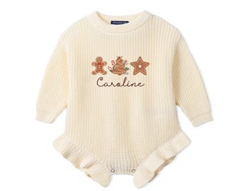 May include: A cream-colored long-sleeve knit romper featuring a ribbed texture, ruffled leg openings, and a central graphic of a gingerbread man, a decorated tree, and a star, with the name Caroline embroidered in brown script lettering below.