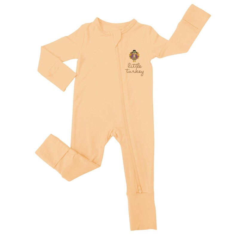 May include: A light peach-colored baby sleeper with long sleeves and attached feet. The sleeper features a full-length zipper and a small graphic of a turkey with the words "little turkey" printed below it. The garment is made of a soft, comfortable material.