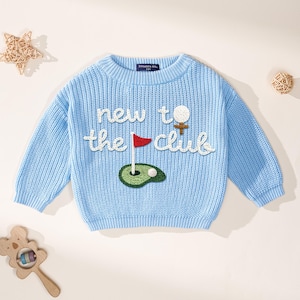 K&ouml;nnte beinhalten: Hellblauer Strickpullover mit dem Schriftzug "new to the club" in wei&szlig;er Schrift. Der Pullover hat ein Golf-Motiv mit Golfball, Abschlag, Fahne und Gr&uuml;n. Baby- oder Kleinkindgr&ouml;&szlig;e.