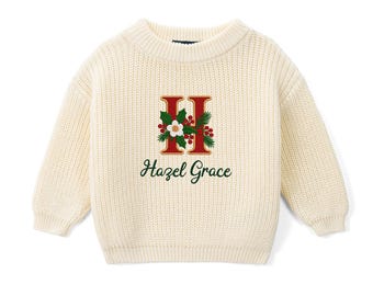 May include: A cream-colored long-sleeve knit sweater featuring a large red letter H decorated with green holly leaves, red berries, and a white flower. Below the letter, the name Hazel Grace is printed in a dark green cursive script.