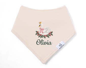 May include: A cream-colored triangular baby bib featuring an embroidered white swan wearing a gold crown and a pink bow. The swan is surrounded by green pine branches and red berries. The name Olivia is embroidered in dark green below the swan.
