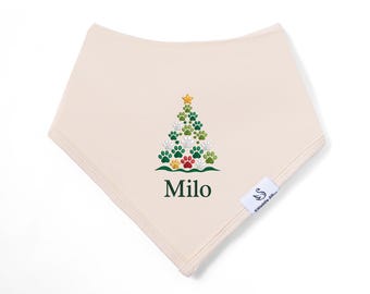 May include: A cream-colored triangular baby bib featuring an embroidered Christmas tree made of green, white, and gold paw prints topped with a gold star. Below the tree, the name Milo is printed in dark green text. A small white tag is attached.