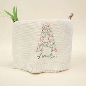 May include: Cream-colored knitted blanket with a floral embroidered letter "A" in pink, blue, and green. The name "Amelia" is embroidered below the letter in green script. The blanket has a ribbed edge.