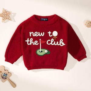 K&ouml;nnte beinhalten: Ein roter Strickpullover mit dem Text "new to the club" in Wei&szlig;. Der Pullover hat ein gesticktes Golf-Motiv, einschlie&szlig;lich Golfball, Fahne und Gr&uuml;n. Der Pullover hat lange &Auml;rmel und einen runden Ausschnitt.