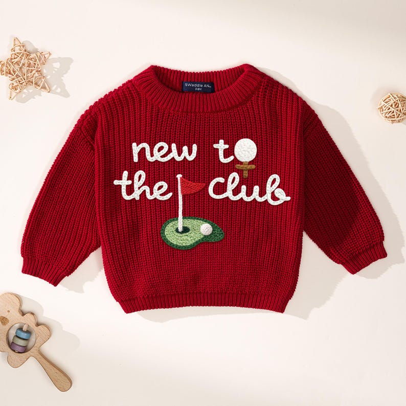 K&ouml;nnte beinhalten: Ein roter Strickpullover mit dem Text "new to the club" in Wei&szlig;. Der Pullover hat ein gesticktes Golf-Motiv, einschlie&szlig;lich Golfball, Fahne und Gr&uuml;n. Der Pullover hat lange &Auml;rmel und einen runden Ausschnitt.