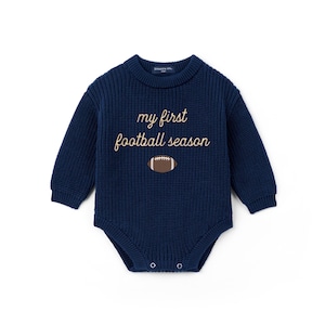 May include: A navy blue long-sleeve knit bodysuit featuring a crew neckline and ribbed texture. The chest displays the embroidered text "my first football season" in a light tan script font, positioned above a small brown and white football graphic.