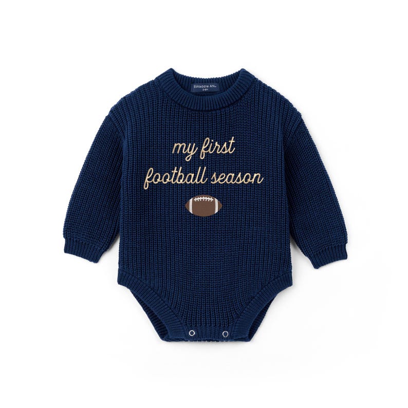 May include: A navy blue long-sleeve knit bodysuit featuring a crew neckline and ribbed texture. The chest displays the embroidered text "my first football season" in a light tan script font, positioned above a small brown and white football graphic.
