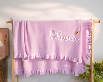 May include: A light pink knitted blanket with a ruffled edge, embroidered with the name "Olivia" in white script. A small yellow butterfly is embroidered near the name. The blanket is displayed on a wooden rack.