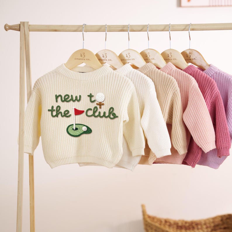 K&ouml;nnte beinhalten: Eine Reihe von Kinderpullovern in verschiedenen Pastellfarben, die an Holzkleiderb&uuml;geln h&auml;ngen. Der vordere Pullover ist cremefarben und tr&auml;gt den Text "new to the club" sowie ein Golf-Motiv.