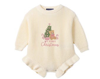 May include: A cream-colored long-sleeve knit romper featuring a ribbed texture and ruffled leg openings. The center displays an embroidered graphic of a teddy bear, a Christmas tree, and candy canes with the text My First Christmas.