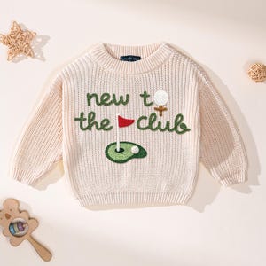 K&ouml;nnte beinhalten: Cremefarbener Strickpullover mit dem Text "new to the club" in Gr&uuml;n gestickt. Das Design umfasst einen Golfball, eine Fahne und ein Gr&uuml;n. Der Pullover hat einen Rundhalsausschnitt und lange &Auml;rmel.