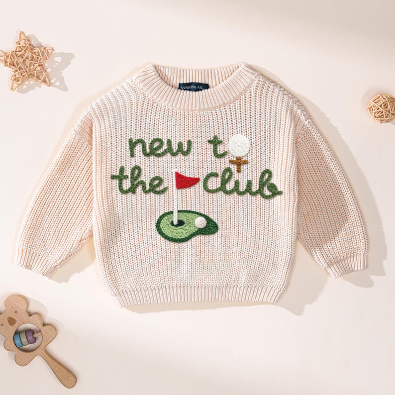 K&ouml;nnte beinhalten: Cremefarbener Strickpullover mit dem Text "new to the club" in Gr&uuml;n gestickt. Das Design umfasst einen Golfball, eine Fahne und ein Gr&uuml;n. Der Pullover hat einen Rundhalsausschnitt und lange &Auml;rmel.