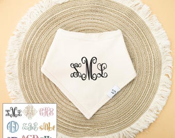 May include: Cream-colored baby bib with a black monogram of the letters SML. The bib is triangular and rests on a round, woven placemat with a fringed edge. A small square tag is attached to the bib. A selection of monogram styles is shown.