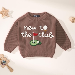 K&ouml;nnte beinhalten: Ein brauner Strickpullover mit dem Text "new to the club" in Wei&szlig;. Der Pullover hat ein Golf-Motiv mit Golfball, Abschlag, Fahne und Gr&uuml;n. Der Pullover hat lange &Auml;rmel und einen gerippten Ausschnitt.
