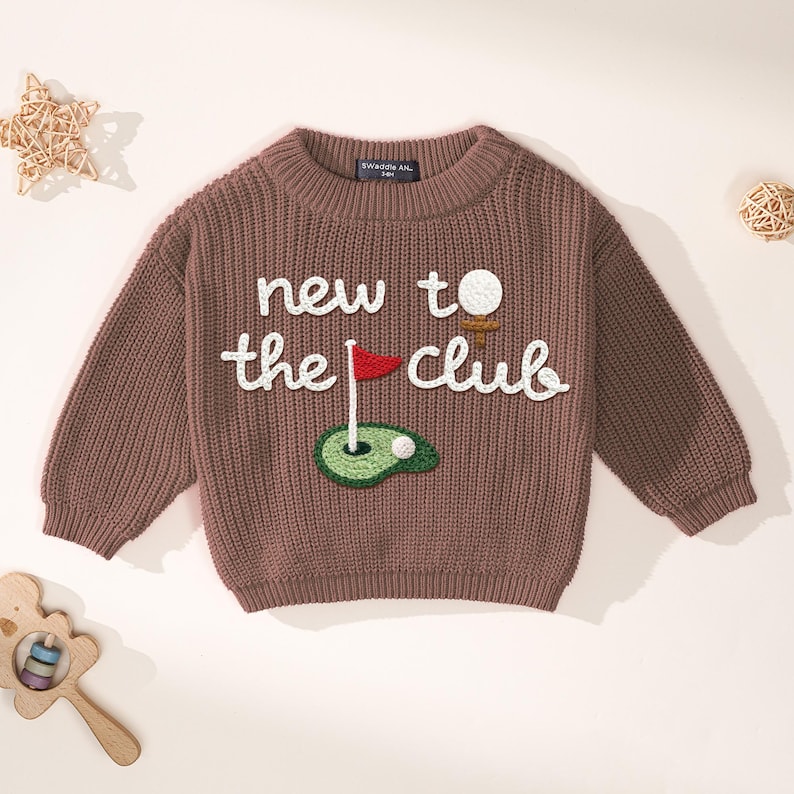 K&ouml;nnte beinhalten: Ein brauner Strickpullover mit dem Text "new to the club" in Wei&szlig;. Der Pullover hat ein Golf-Motiv mit Golfball, Abschlag, Fahne und Gr&uuml;n. Der Pullover hat lange &Auml;rmel und einen gerippten Ausschnitt.