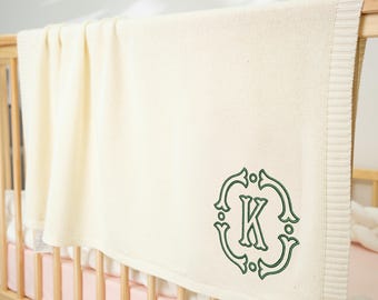 May include: Cream-colored baby blanket with a green embroidered initial "K" in a decorative frame. The blanket is draped over a wooden crib, with a pink sheet visible underneath. The blanket has a knitted texture.