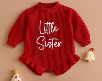 May include: A red knitted baby romper with the words "Little Sister" in white script. The romper has long sleeves, a ruffled hem, and snap closures. A small white Christmas tree and a bell ornament are next to the romper.